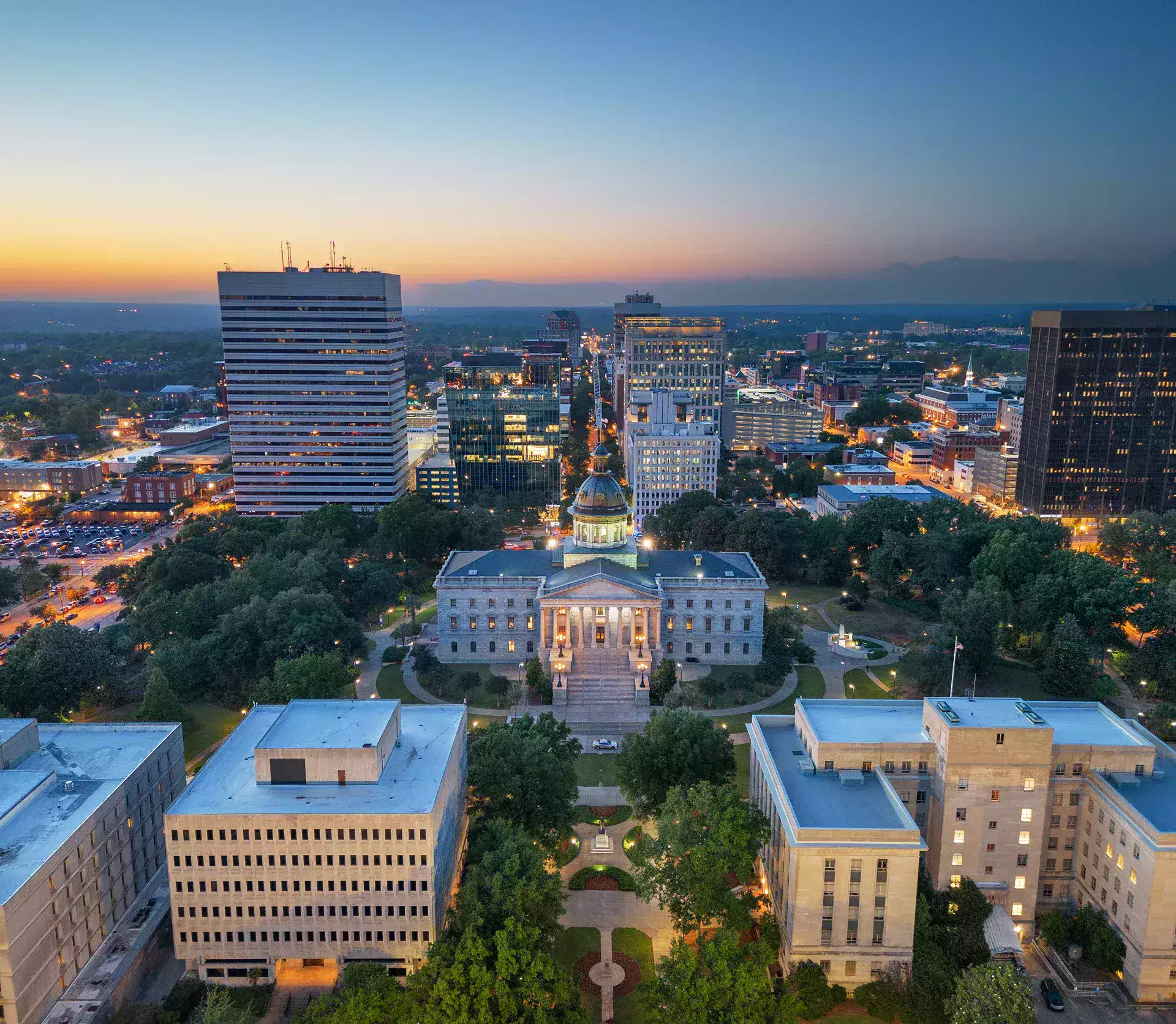 Skyline view of a city at dusk, featuring a historic building with a dome, surrounded by modern skyscrapers and green parks.