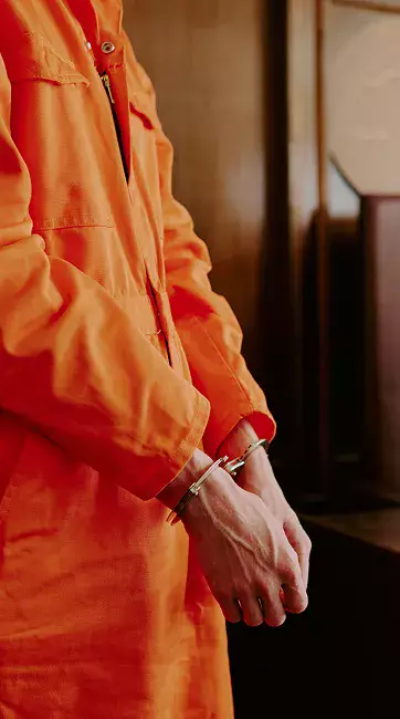 A person wearing an orange jumpsuit stands with their hands clasped, showing silver handcuffs against a wooden background.