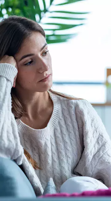 A person wearing a cozy, knitted sweater sits on a couch, looking thoughtfully toward a window, with green plants in the background.