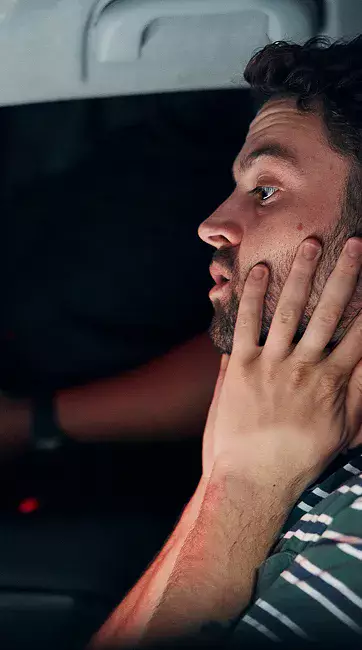 A person wearing a striped shirt is sitting in a car, resting their chin on their hand, with a blurred face and a dark background.