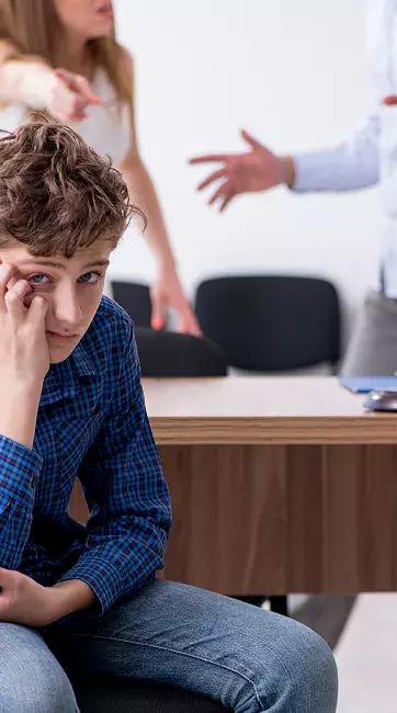 A young person sits with their head down in a classroom setting, while adults in the background argue, showing a tense atmosphere.