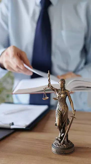 A bronze statue of Lady Justice stands on a desk while a person reviews a book, symbolizing law and justice.
